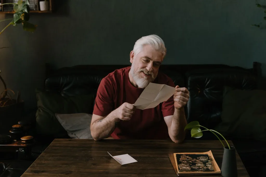 An older man with white hair reading a letter at a wooden table at home